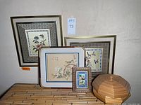 Overall view of four Asian art pieces: two embroidered silk floral art pieces, one Chinese silk painting, and a small painted glass tile with detailed frame, photographed against bamboo surface.