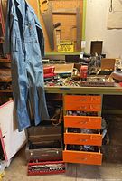 Wide view showing blue Ben Davis work overalls hanging, several orange drawers filled with tools, metal toolboxes partially open revealing hand tools on bench.