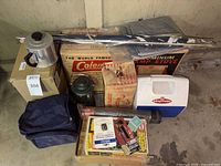 Overview of camp gear including Coleman aluminum camp stove box, green Coleman lantern, battery operated lantern, Playmate cooler, soft cooler bag, and assorted accessories on the floor.