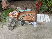 Overview of all lot contents arranged on concrete surface including metal dragonflies, ceramic bird figurines, terra-cotta cat, glass votive holder, ceramic flower relief tiles.