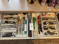 Photo of two white organizer trays containing stainless steel forks, spoons, knives, and various kitchen utensils including bottle opener, silicone brush, vegetable peeler, can opener, and manual kitchen tools with Wusthof knife sharpener visible on the left tray.