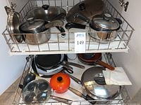 Upper cupboard shelf holding assorted pots and pans including saucepans with lids, frying pans, and an orange LeCreuset small pot with lid.