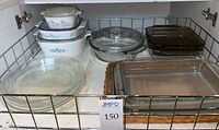 Wide view of assorted baking dishes including clear glass pie plates, rectangular dishes, ceramic casserole, and Corning glass baking dishes in cabinet shelf