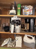 Shelf with assorted small appliances including Cuisinart Mini Prep Processor, Donvier Ice Cream Maker box, Black & Decker coffee maker with glass carafe, Oster can opener, Hamilton Beach hand mixer, and two white irons. Box of cone coffee filters also visible.