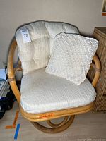 Swivel cane chair with off-white textured foam cushions and an additional crocheted cushion placed on the seat. Shows some signs of wear and staining on fabric.