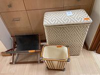 Photo showing the front view of the wicker hamper, wooden trashcan, and sling magazine holder together on a wooden floor near a cabinet.