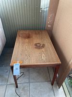 Full view of the wood side table showing rectangular top with visible water stains and fading, alongside a chair and wall background.