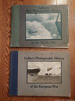 Two hardcover antique books laid flat on wood floor, showing front covers with war-related photographs and titles.