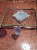 Photo showing clear quartz pyramid with visible corner chip and purple fluorite crystal mounted on a gold-tone frame stand placed on wood surface.