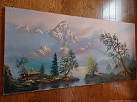 Front view of large oil painting showing mountain, lake, trees, and cabin in landscape scene with signature visible in lower right.