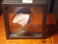 Lepidolite mineral specimen displayed in a black-framed case on wooden surface under lighting.