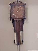 Front view of the weighted pendulum Urgos wall clock mounted on a wall, showing clock face and weights.