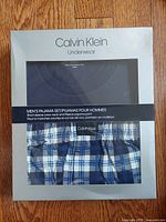 Front view of Calvin Klein branded box showing navy short sleeve crew neck top and plaid pajama pants in blue, white, and black.