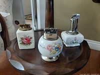 View of three vintage ceramic lighters with floral hand painted designs on a wooden table