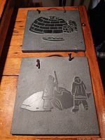 Two square soapstone plaques showing Inuit relief carvings; top plaque depicts whale hunt with two Inuit figures and whale; bottom plaque depicts igloo build with igloo and figure.