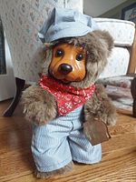 Front view of Robert Raikes Bears wood face bear wearing blue and white striped overalls, matching cap, and red bandana around neck. Bear has a carved wooden face with black eyes, brown nose, and brown plush fur.