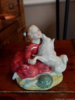 Front angle view of the Royal Doulton Home Again figurine showing the girl in a red dress embracing a white dog, sitting on a green base with a hat detail.