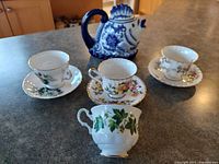 Five vintage to antique fine china teacups on saucers with floral patterns and an ivy leaf design teacup in front, placed on a wooden surface