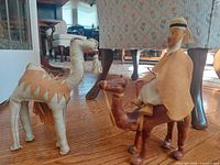 Photo shows both figurines, camel rider mounted on camel, with pack camel beside them on wooden floor indoors.