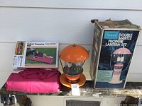 Photo showing Sears propane lantern set with box, orange lantern, and pink rolled fabric camping mat next to it.