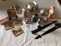 Overview of the ceramic collectibles lot showing containers, coasters, pottery clogs, small figurines, and iron candle straps on a white textured tablecloth.