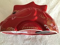 Red ceramic dragon coin bank laid on white textured fabric showing top and side views with decorative gold and white swirl patterns and dragon details.