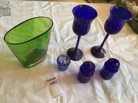 Photo of two tall cobalt blue glass candelabras, two matching cobalt blue goblets, green glass vase, and three small cobalt blue glass items on a white cloth background