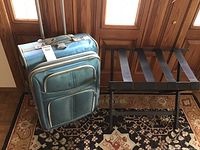 Light blue Delsey suitcase next to a dark brown wooden luggage stand on a patterned rug in front of wooden doors
