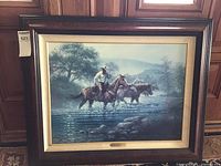 Framed lithograph showing a western scene with horse riders crossing water in a brown frame with gold inner border.