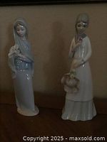 Photo showing two porcelain girl figurines side by side. Left is the Lladro 'Girl with Calla Lilies' with a blue shawl and flowers in hand. Right is the Miquel Requena figurine holding a bonnet and a flower.
