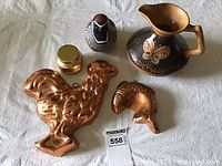 Photo of pottery pitcher with butterfly decoration, two copper gel molds shaped like a rooster and fish head, ceramic quail candle, and small jar with gold lid on white surface.