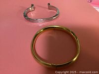 Front view of two bracelets on pink background, one silver open cuff and one gold tone bangle.