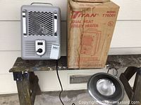Titan dual heat utility heater in gray with protective grille and original box, placed on wooden surface next to trouble light.