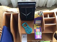 Wooden cubby with compartments containing a black planner, turquoise calculator, Post-it notes, and small office supplies arranged inside.