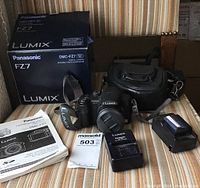 Photo showing Panasonic Lumix DMC-FZ7 camera with accessories including original box, manuals, a carrying case, battery charger, camera strap, and a MaxSold tag on an upholstered chair.