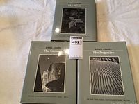Three Ansel Adams hardcover photography books titled The Camera, The Negative, The Print laid out on a white surface showing their front covers with photographic art.
