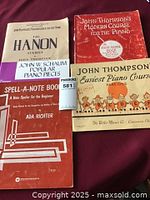 Four beginning piano sheet music books shown on blanket: The Hanon Studies, John Thompson's Modern Course for the Piano, John Thompson's Easiest Piano Course, and Spell-A-Note Book by Ada Richter.