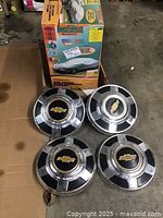 Four Chevrolet hubcaps displayed on cardboard, showing chrome and black colors with gold Chevy logos.