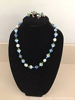 Front view of necklace and matching clip earrings displayed on black velvet stand. Shows arrangement of blue-green art glass beads, faux pearls.
