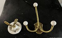 Two vintage-style coat hooks: one double hook with white ceramic base and brass arms, one triple brass hook with white ceramic knobs laid on black surface.