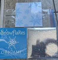 Origami snowflake kit contents laid out on tiled surface. Includes instruction booklet and two types of decorative origami paper.