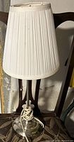 Table lamp with white pleated shade on chair, showing lampshade details, base, pull chain, and attached power cord.