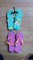 Photo showing two pairs of flip flops on wooden floor. Top pair turquoise with pineapple print and store tag on strap; bottom pair purple with orange straps and floral design.