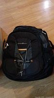 Photo of a black Eddie Bauer backpack placed on a wooden floor, showing front view with multiple compartments and bungee cords.