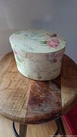 Closed view of the oval floral decorative box sitting on a wooden stool, showing box shape and floral pattern