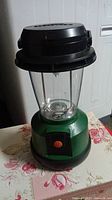Front view of green and black camping lantern showing transparent middle and orange power button