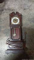 Photo of a working electric mini grandfather clock with wooden case, Roman numeral clock face, brass pendulum, and attached power cord.