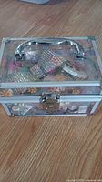Closed clear plastic jewelry box with silver metal frame and latch sitting on wooden floor, showing colorful flower design on sides and handle on top.