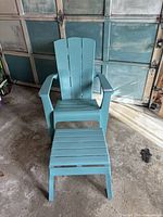 Front view of plastic Adirondack patio chair with ottoman in garage setting, showing chair's wide armrests and curved back design with slats.