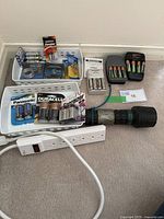 View of assorted batteries in and out of packaging, white plastic baskets, power strip, and flashlight on floor.